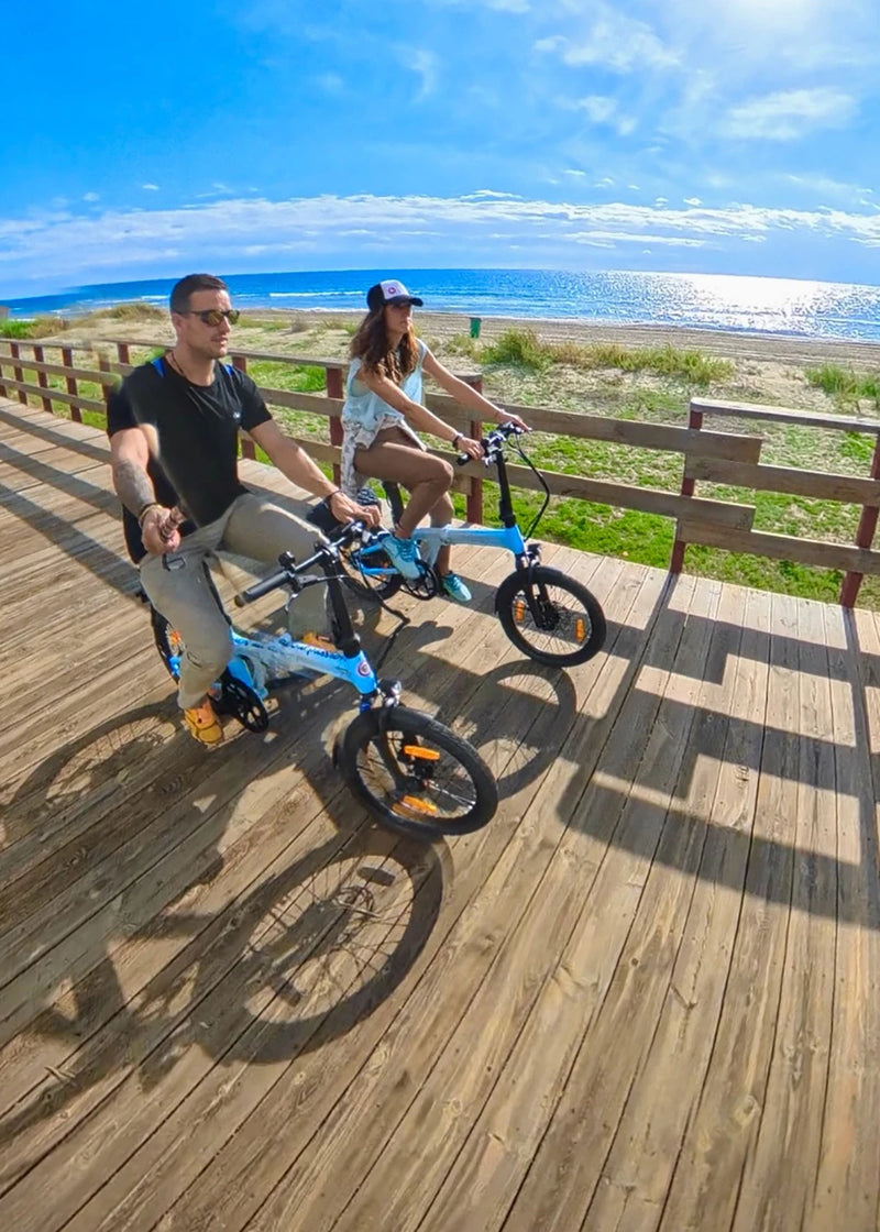 Couple riding blue folding ebikes on a coastal boardwalk.