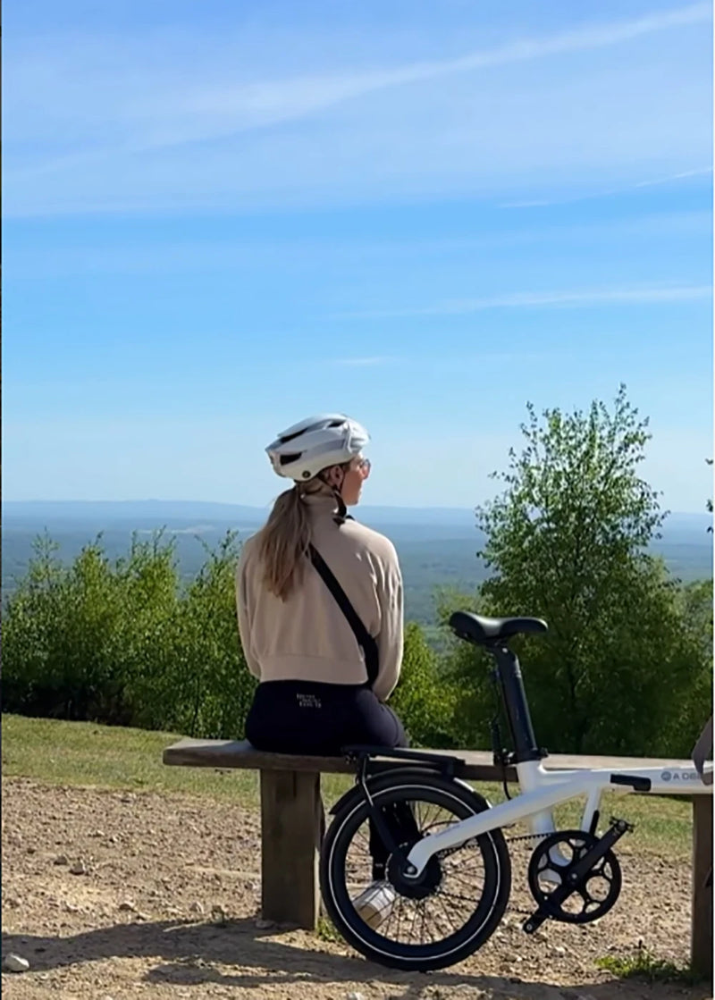 Woman with a white folding ebike sitting on a bench overlooking a valley.