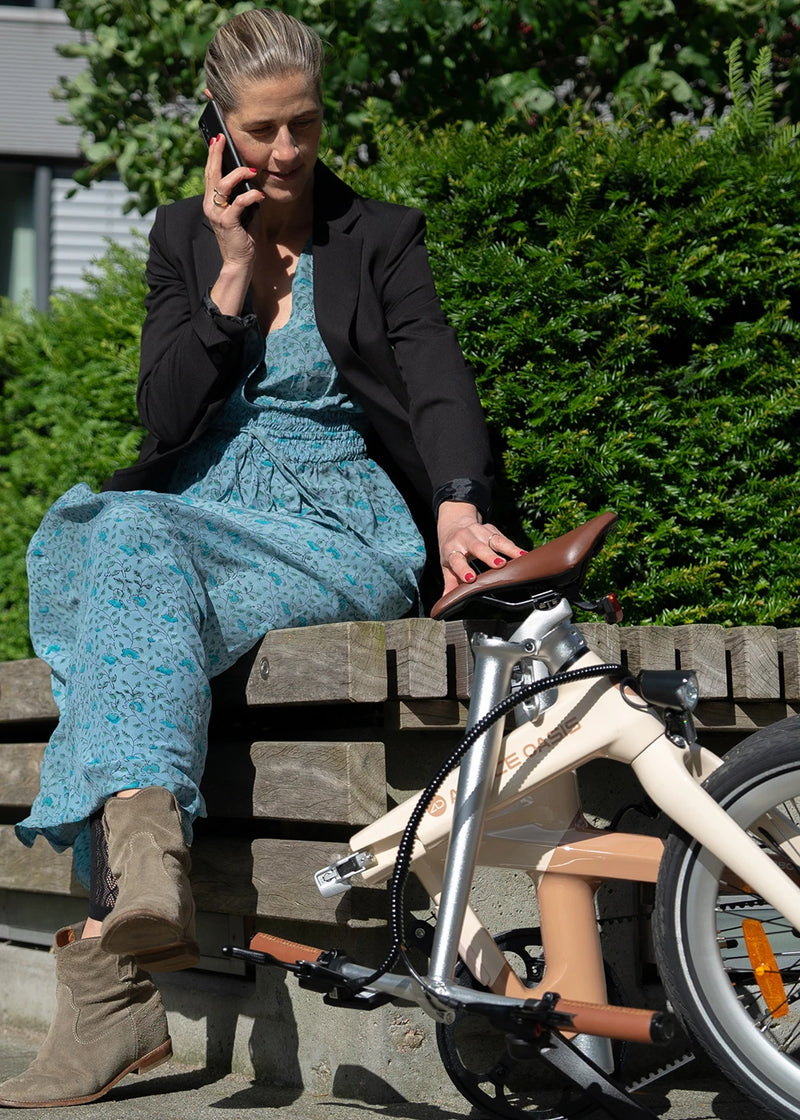 Woman in a dress and blazer talking on the phone next to a cream folding ebike on a bench.