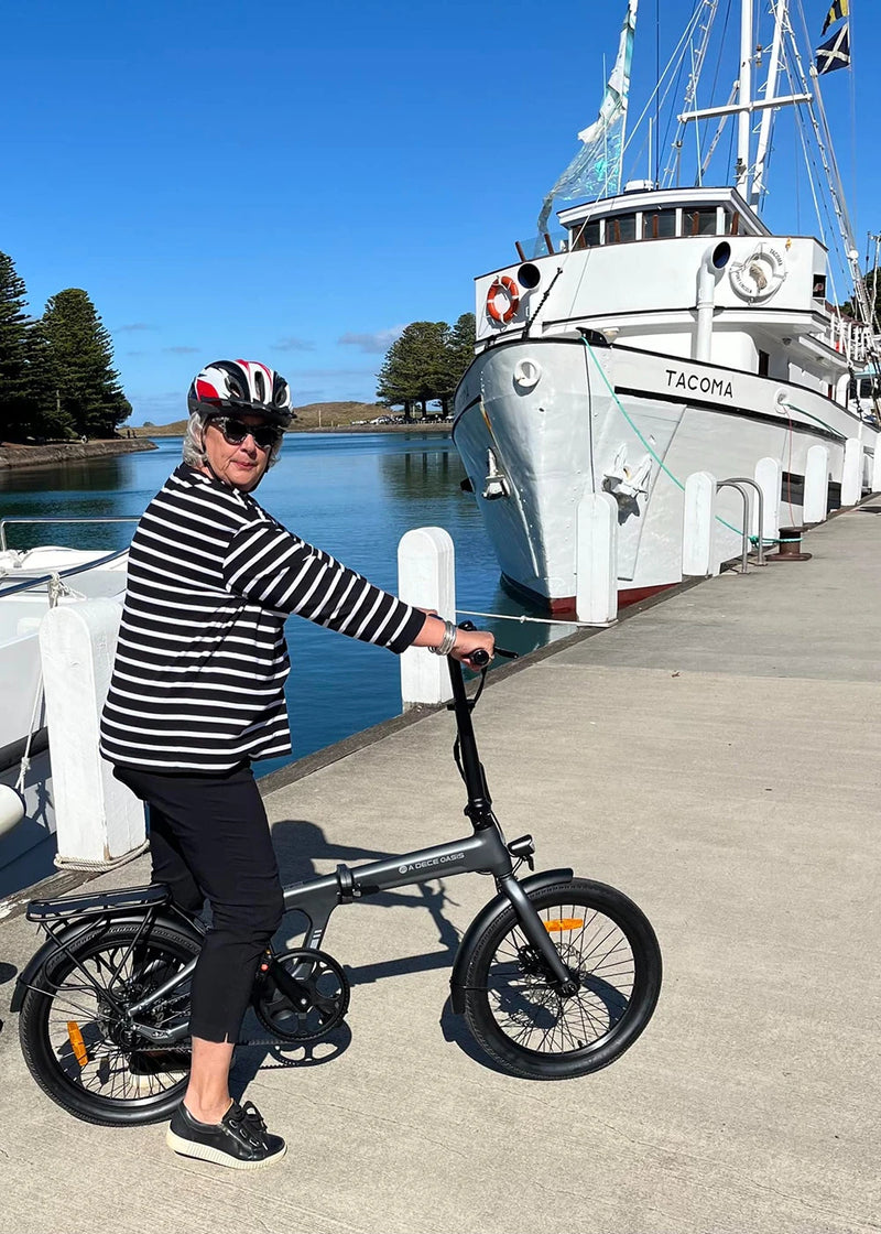Woman in a helmet standing with a dark grey folding ebike on a dock next to a ship.